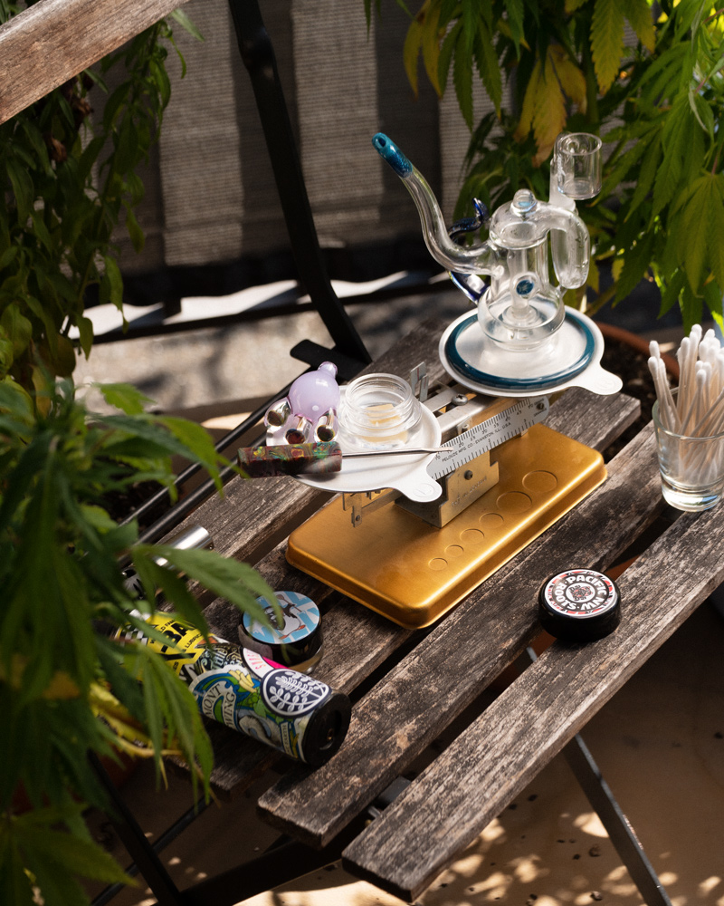 A complete dabbing setup and a triple-beam scale sit on an outdoor table, surrounded by cannabis plants.