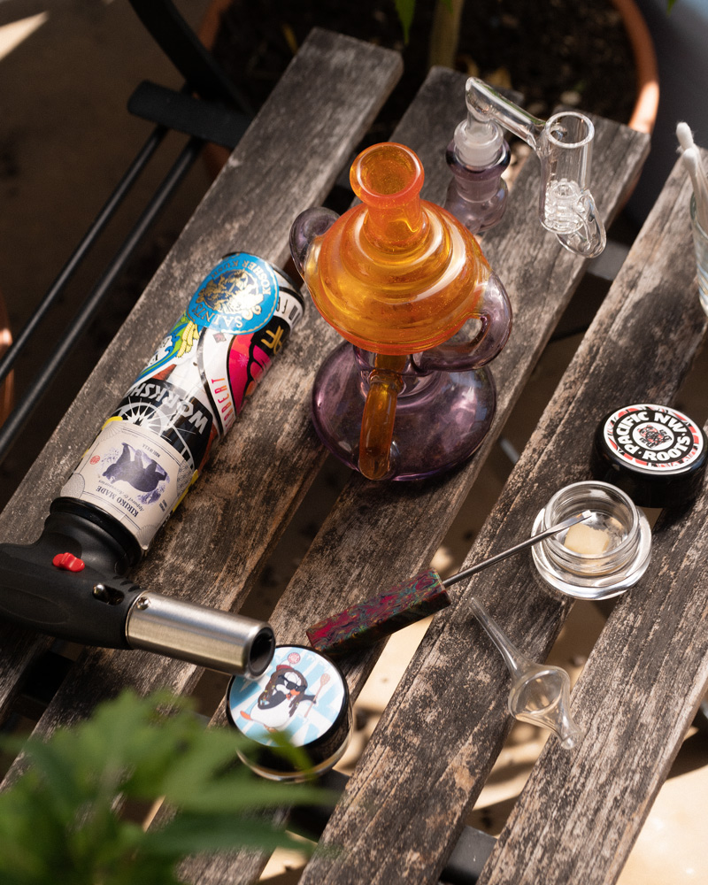 A recycler-style dab rig, torch, carb cap, and several grams of cannabis concentrate sit on an outdoor table. 