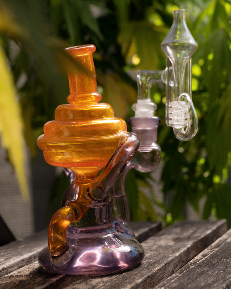 A recycler-style dab rig with a carb cap sits on an outdoor table. Cannabis plants are visible in the background.