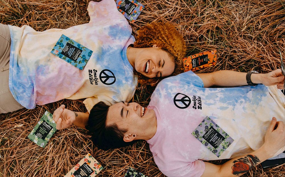 Two people laughing and laying in opposite directions on hay, surrounded by Smokiez Edibles